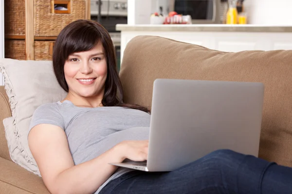 Mujer embarazada en el sofá con ordenador portátil —  Fotos de Stock