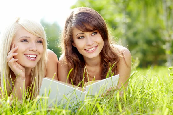 Två unga vackra leende kvinnor läser bok, sitter på gräset — Stockfoto
