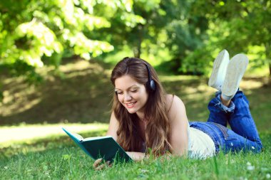 Young woman with headset reading book in park clipart