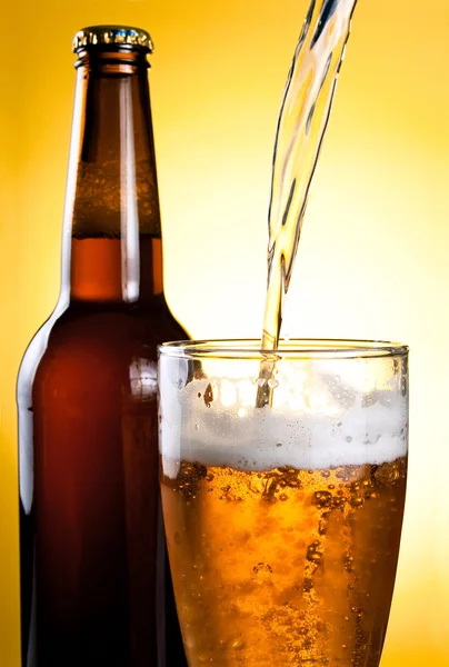 Cerveja sendo derramada em vidro e garrafa no fundo amarelo — Fotografia de Stock