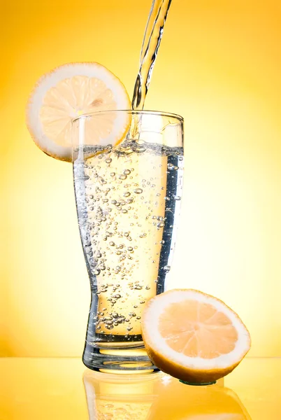 Pouring of mineral water in glass with a lemon on a yellow backg — Stock Photo, Image