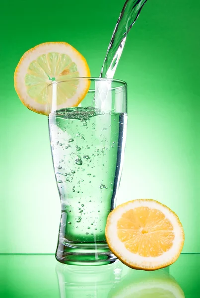 Pouring of mineral water in glass with a lemon on a green backgr — Stock Photo, Image