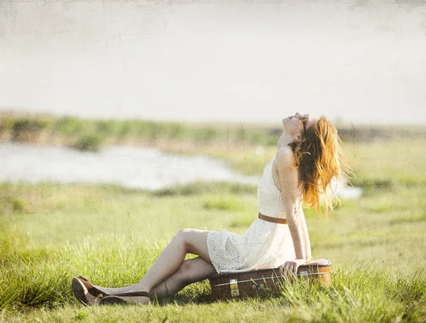 Ung flicka med resväska sitter på våren gräs nära lake. — Stockfoto