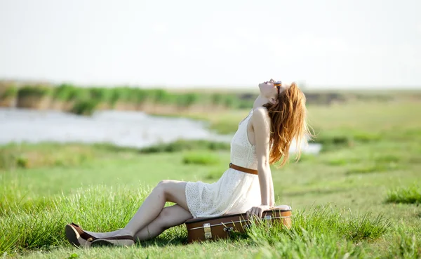 Jonge mode meisje met koffer zittend op lente gras in de buurt van lake. — Stockfoto