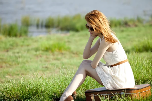 Junges Modemädchen mit Koffer sitzt am Frühlingsgras am See. — Stockfoto