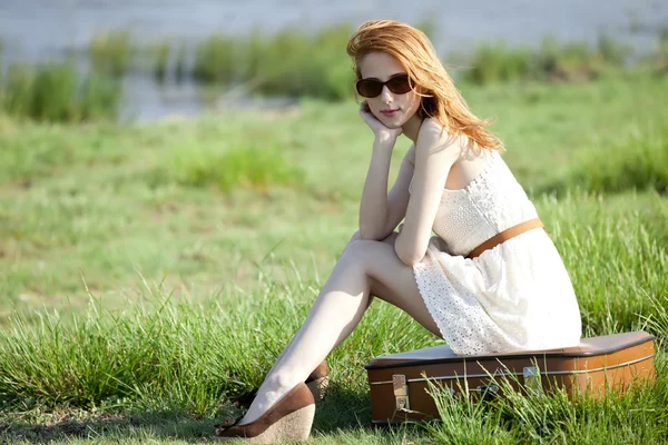 Young fashion girl with suitcase sitting at spring grass near lake. — Stock Photo, Image