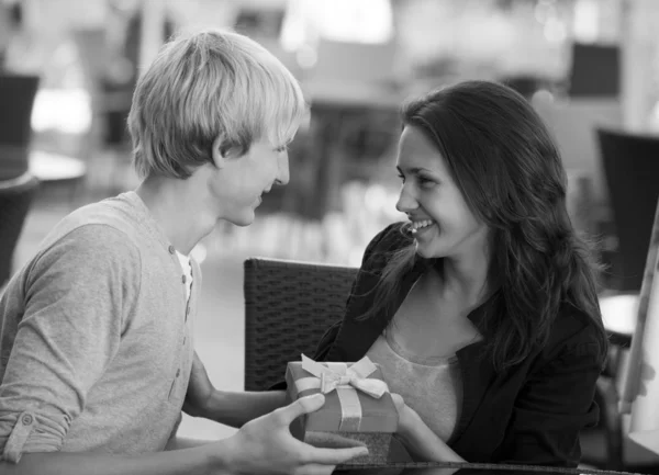 The young man gives a gift to a young girl in the cafe and they — Stock Photo, Image