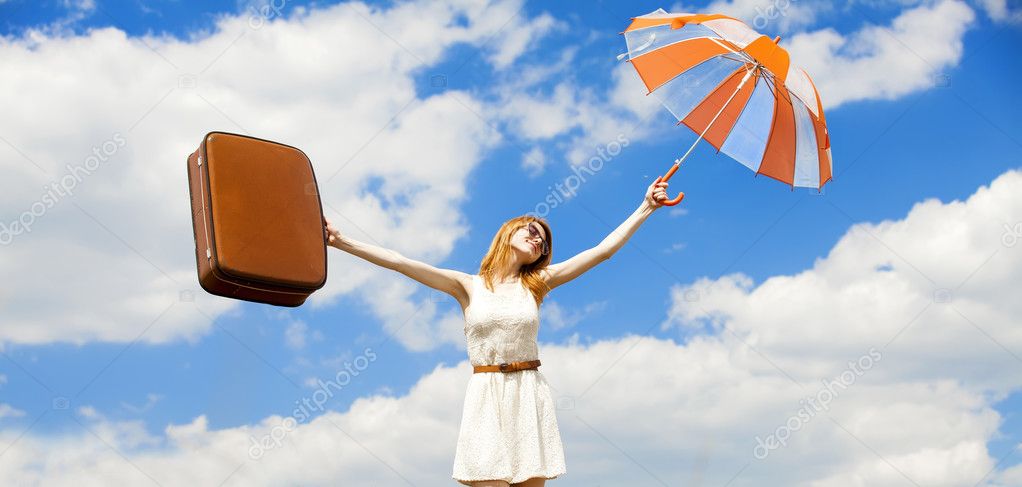 Beautiful redhead girl with umbrella and suitcase at blue sky ba