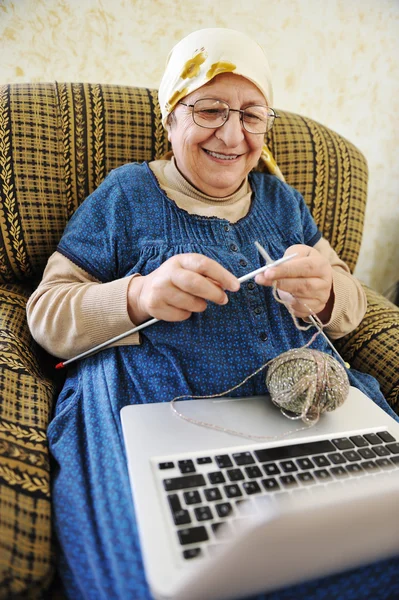 Elderly woman working on laptop in house and knitting — Stock Photo, Image
