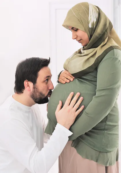 怀孕的穆斯林妻子与丈夫亲吻的肚子和他的手在肚子上微笑 — 图库照片