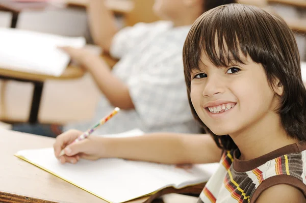Bonito menino positivo escrevendo e sorrindo. Interior, escola, sala de aula . — Fotografia de Stock