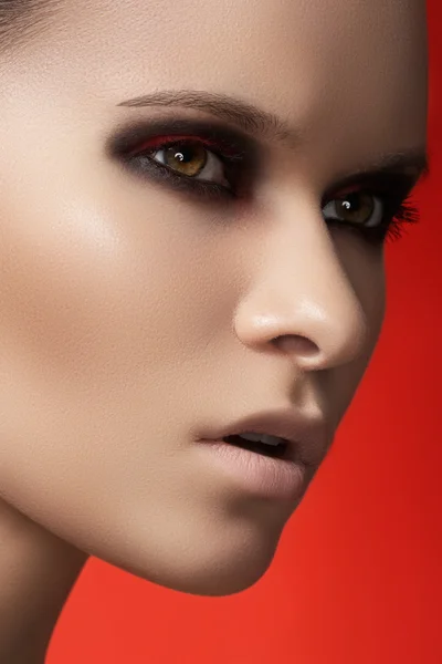 Retrato close-up de belo modelo de mulher jovem no fundo vermelho brilhante. Moda escura maquilagem de olhos fumegantes, lábios pálidos e pele lisa — Fotografia de Stock