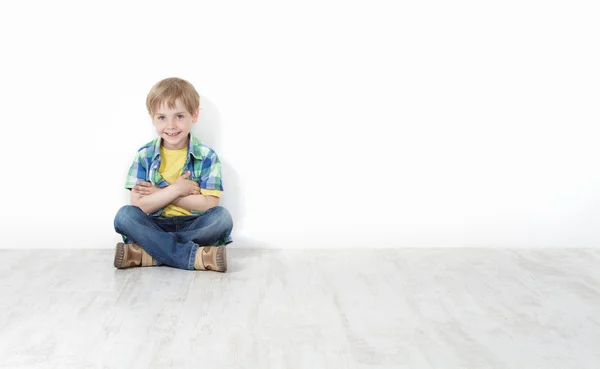Красивый маленький мальчик, сидящий на полу, прислонившись к белой стене — стоковое фото