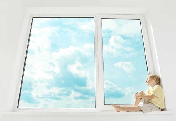 เด็กนั่งอยู่บนระเบียงหน้าต่างและฝัน หน้าต่างบานใหญ่และท้องฟ้า . — ภาพถ่ายสต็อก