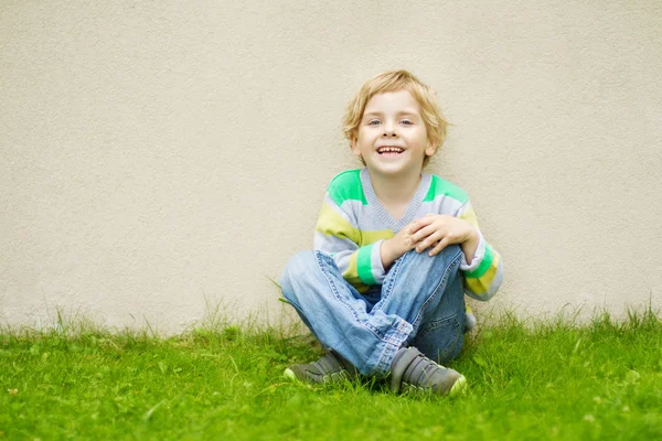 Улыбающийся мальчик, сидящий на траве у бетонной стены — стоковое фото