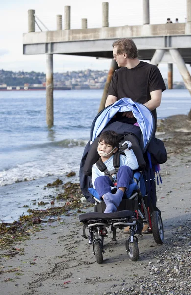 Отец толкает инвалидное кресло с сыном-инвалидом на пляже — стоковое фото