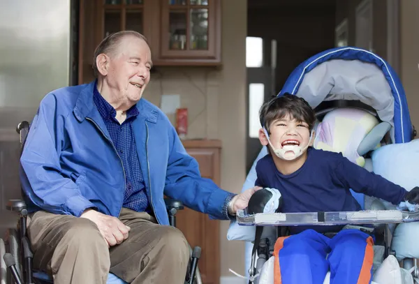 Пожилой человек в инвалидной коляске смеется с мальчиком-инвалидом на кухне — стоковое фото