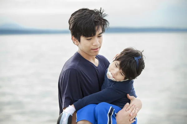 Big brother carrying disabled boy by lake shore — Stock Photo, Image