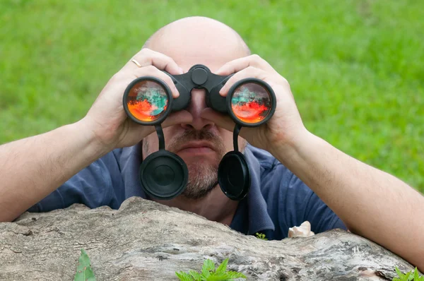 Bald man watching the nature through binoculars while leaning on old tree