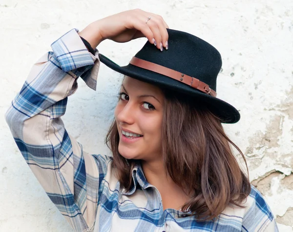 Beautiful cowgirl smiles while holding her hat and leaning on the old wall — Stock Photo, Image