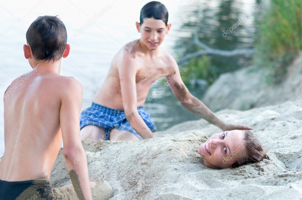 Two teenage boys covering with sand teenage girl on river beach