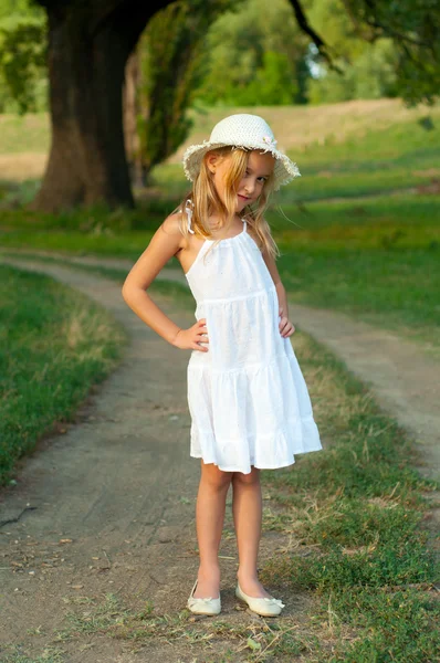 Roztomilé děvčátko v bílých šatech a klobouk, stojící na zemi silnici na krásné letní den — Stock fotografie