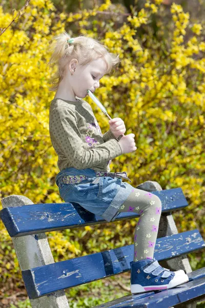 봄에 벤치에 앉아 막대 사탕과 어린 소녀 — 스톡 사진
