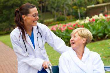 Friendly female doctor pushing senior patient in wheelchair outdoors clipart