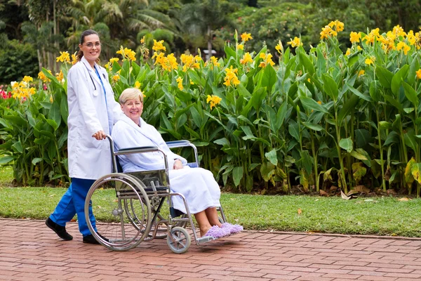 Cuidar médico fêmea empurrar paciente sênior na cadeira de rodas ao ar livre — Fotografia de Stock