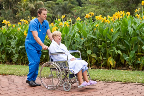 Enfermera médica masculina empujando a paciente mayor en silla de ruedas al aire libre —  Fotos de Stock