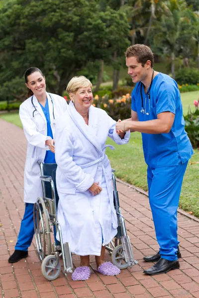 Young caring doctor and nurse helping senior patient get up from wheelchair — Stock Photo, Image