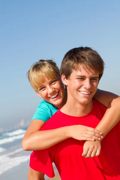 Teen brother and sister piggyback on beach — Stock Photo, Image