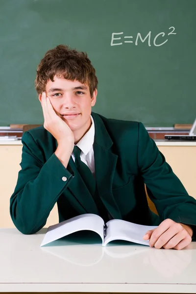 Menino do ensino médio sorrindo com livro na mesa em sala de aula — Fotografia de Stock