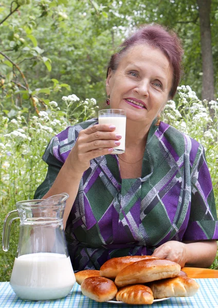 The happy elderly woman eats pies with fresh milk — Stock Photo, Image