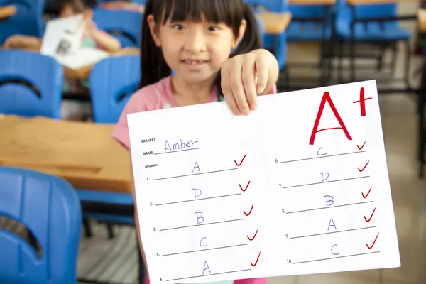 Smiling little girl showing exam paper with a plus in the classroom — Stock Photo, Image