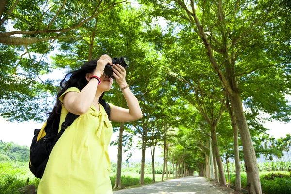 Young woman with backpack standing in the green forest taking photo — Stock Photo, Image