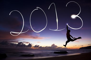 Happy new year 2013. young man jumping and drawing 2013 by flashlight in the air on the beach before sunrise clipart