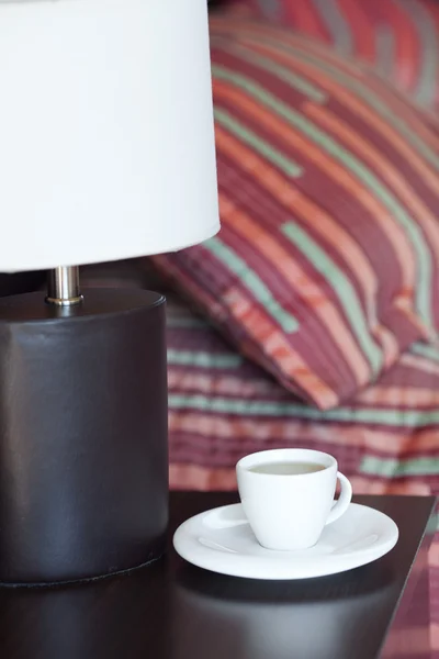 Postel s polštáři, šálek čaje na nočním stolku a lampa — Stock fotografie