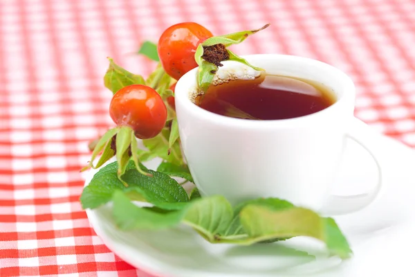 Filiżankę herbaty i owoców dzikiej róży jagody z liści na kratę tkaniny — Zdjęcie stockowe