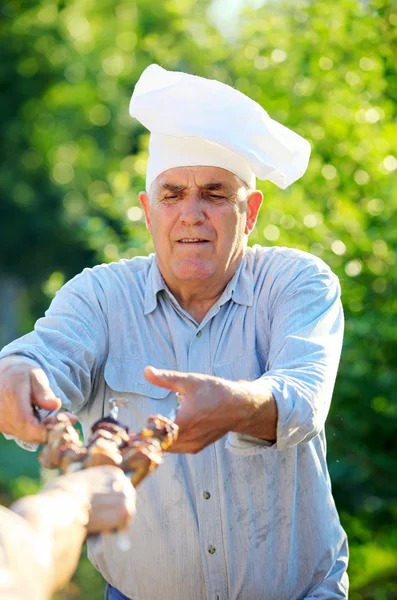 Mature Caucasian man in chef's hat holding grilled shish kebabs