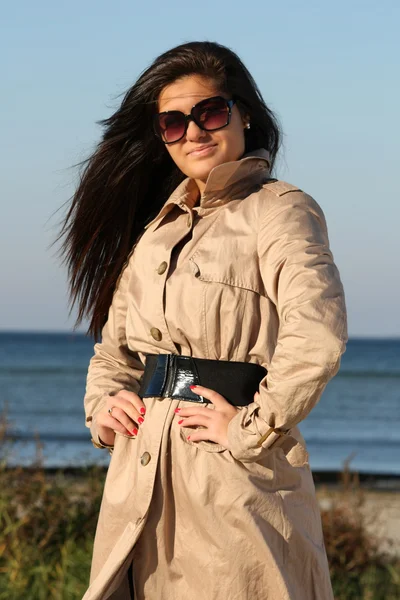 Γυναίκα σε μπεζ φθινόπωρο παλτό και γυαλιά ηλίου που ποζάρει δίπλα στη θάλασσα — Φωτογραφία Αρχείου