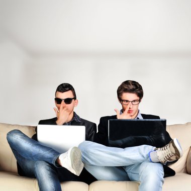 Two young gamers sitting together on sofa and using their laptop clipart