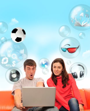 Adult couple sitting on sofa and having fun using their laptop. clipart