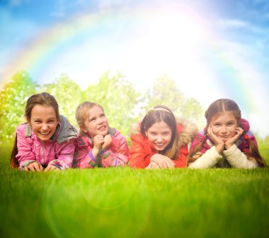 Happy group of girls lying on a green grass. Rainbow and sun ove clipart