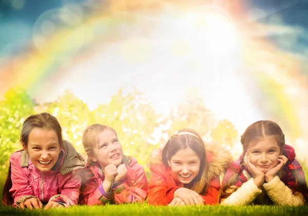 Fröhliche Gruppe von Mädchen, die auf einem grünen Gras liegen. Regenbogen und Sonne — Stockfoto
