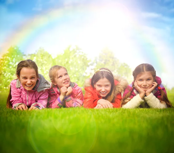 Fröhliche Gruppe von Mädchen, die auf einem grünen Gras liegen. Regenbogen und Sonne — Stockfoto