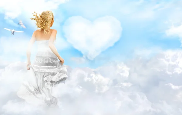 Giovane donna felice che corre attraverso le nuvole nel cielo. Cuore su sfondo — Foto Stock