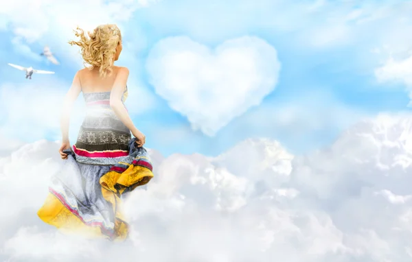Glückliche junge Frau, die über Wolken am Himmel rennt. Herz auf Hintergrund — Stockfoto