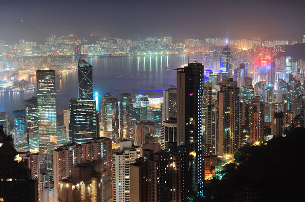Hong Kong city skyline at night with Victoria Harbor and skyscrapers illuminated by lights over water viewed from mountain top.