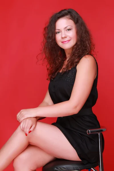 Fiatal nő fekete ruha a divat ülés felett piros háttérre fedett studio Studio képe — Stock Fotó